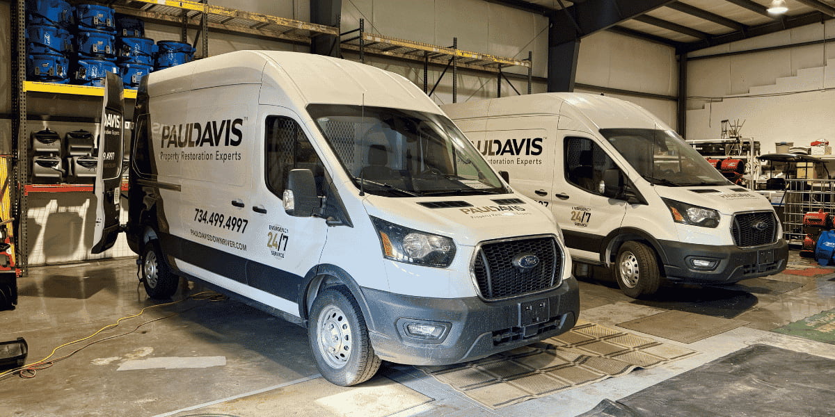 Two white and black Paul Davis Property Restoration Ford vans parked side-by-side inside a brightly lit warehouse facility lined with equipment shelves.